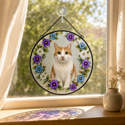 A circular hanging suncatcher with a photo of an orange and white cat surrounded by purple, blue, and white flowers, displayed on a window with sheer curtains and a small vase of dried flowers.