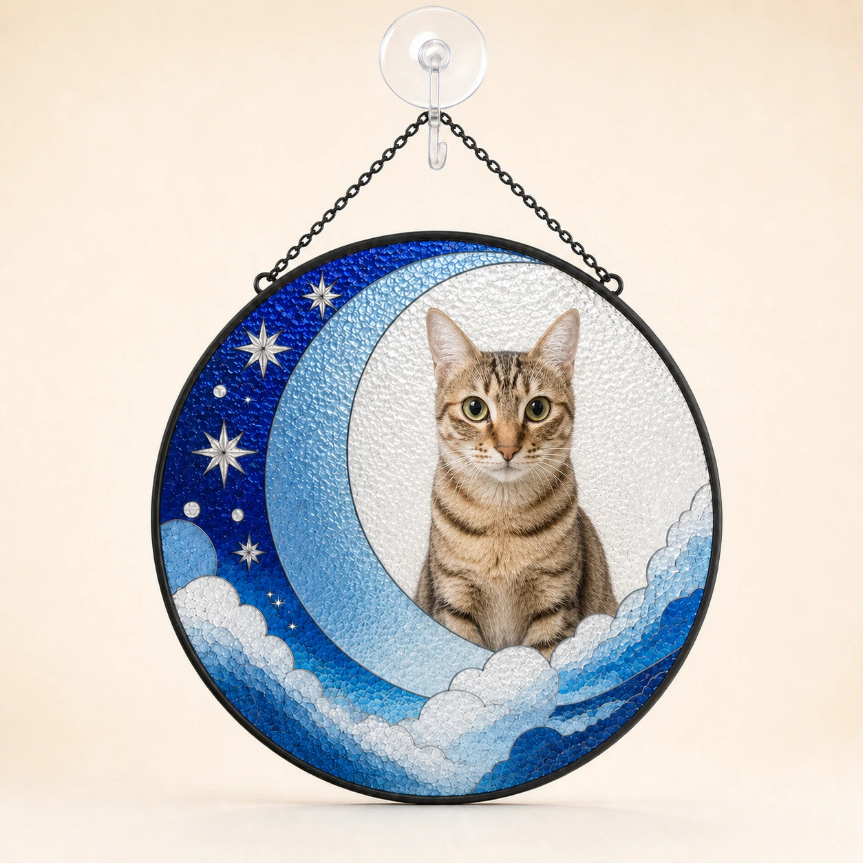 A circular hanging stained-glass-style wall decor piece featuring a tabby cat seated on a crescent moon with blue clouds and white stars, suspended from a clear suction cup with a black chain against a plain beige background.